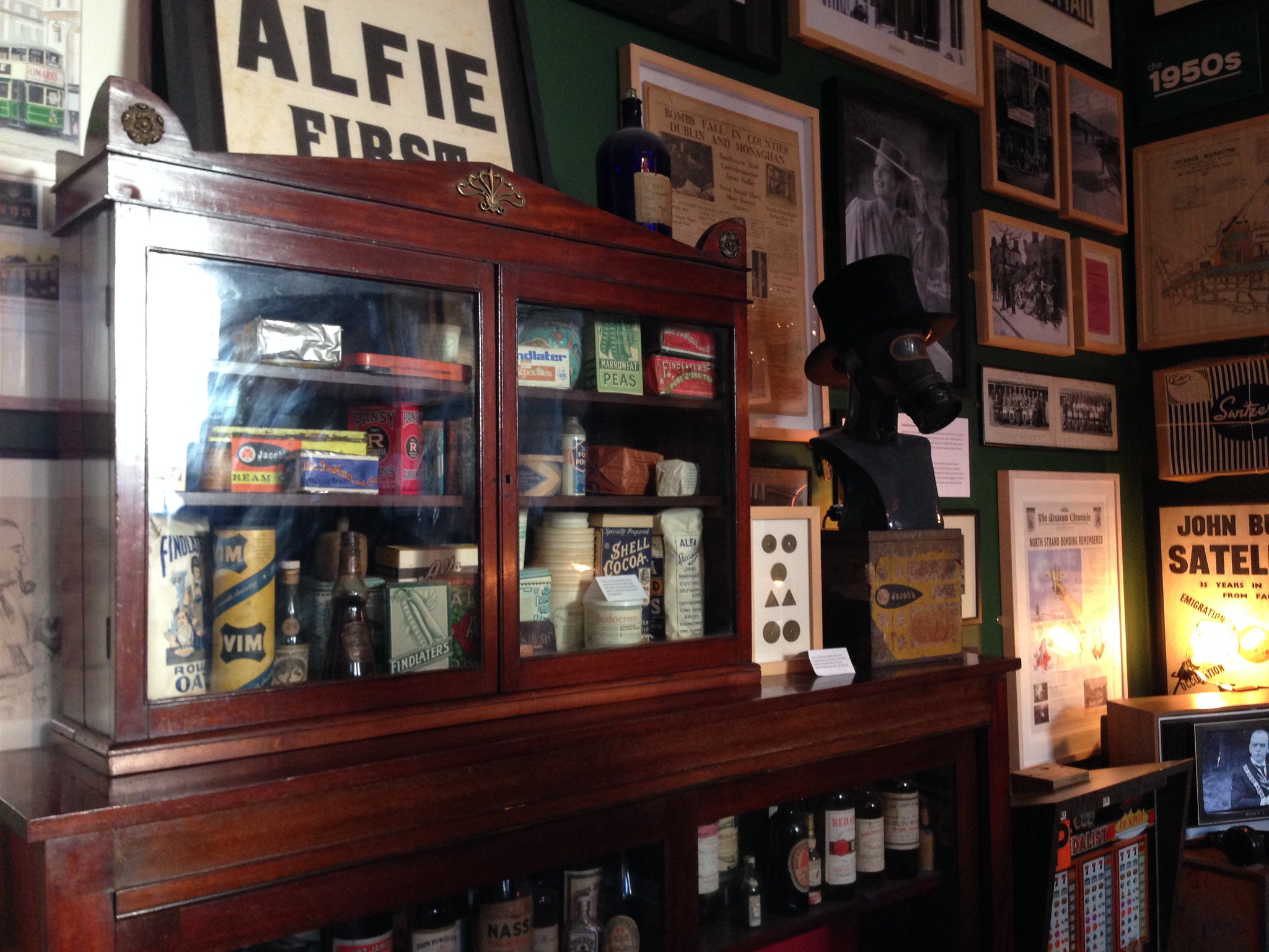 Cabinet filled with antique goods and bottles, gas mask with hat, and framed historical prints at and exhibit in the Little Museum of Dublin, one of the best places to visit in Ireland
