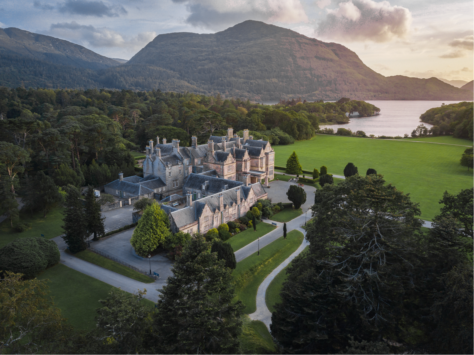Aerial view on the vast Muckross House, one of the best places to visit in Ireland, beside a trimmed green field and a thick forest