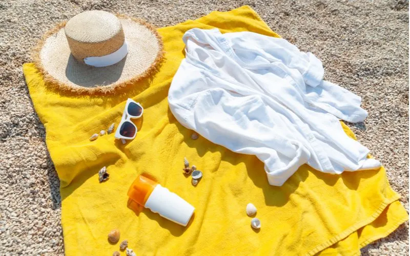 Image for a piece titled Best Beach Blankets featuring a yellow blanket sitting on small pebbles with a summer hat and coverup next to it
