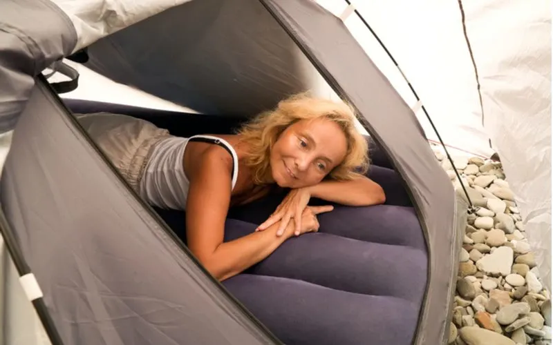 Older woman in a camisole and hiking pants lying down in a tent on the best camping air mattress to pad her sleep from the rocks below