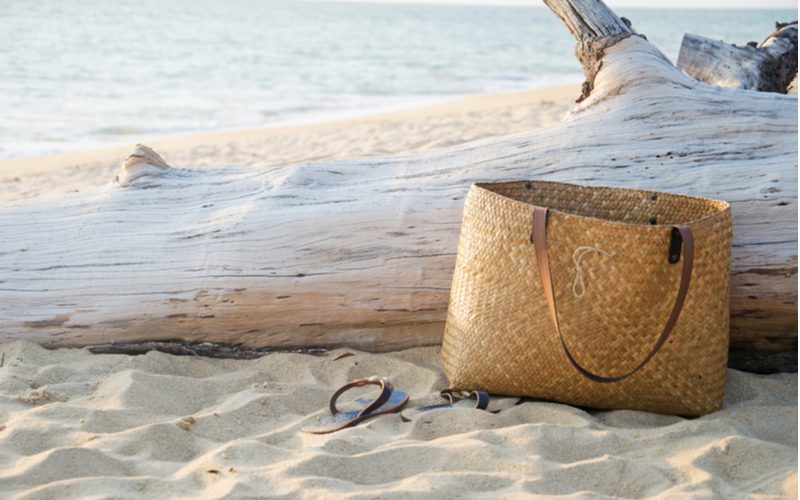 A brown woven beach bag sits on the sand next to flip flops