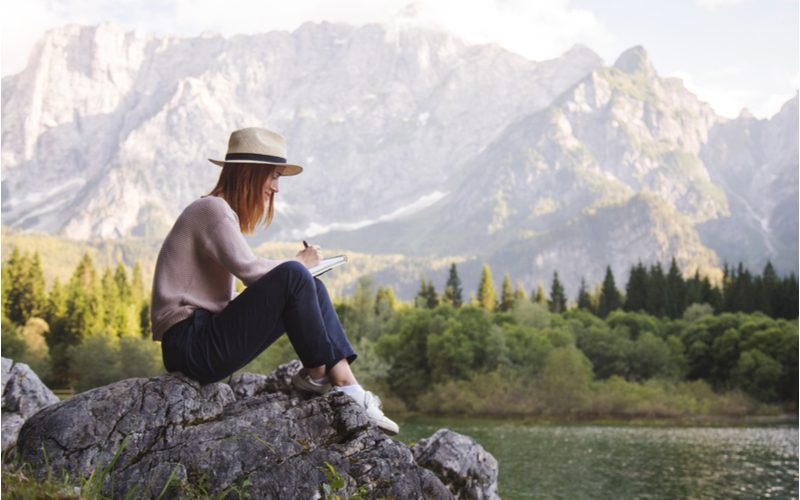 Woman journaling in the best travel journal while sitting on a mountain rock