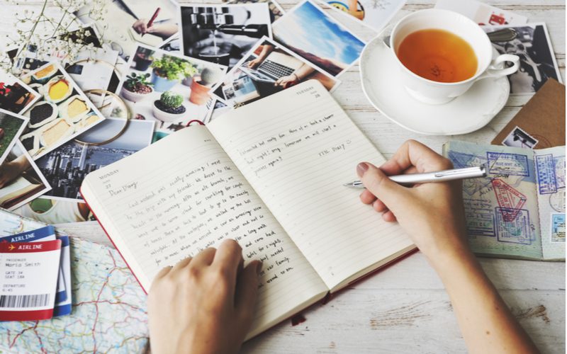 Woman writing in the best travel journal on a table with photographs surrounding it