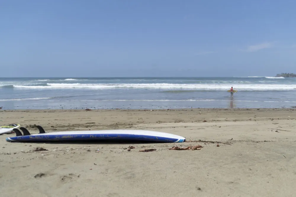A surfboard lying on fine beach and in background, a man carrying his surfboard on a clear day at Playa Iguanita, one of the best beaches in Costa Rica