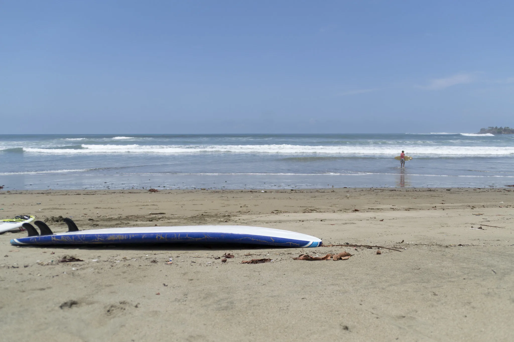 A surfboard lying on fine beach and in background, a man carrying his surfboard on a clear day at Playa Iguanita, one of the best beaches in Costa Rica