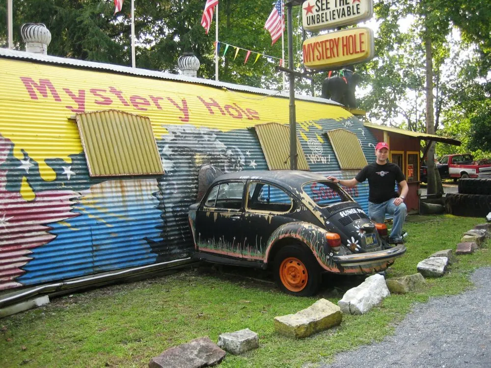 A man posing for a beat up beetle displayed as an attraction at The Mystery Hole, a piece on the best attractions in West Virginia