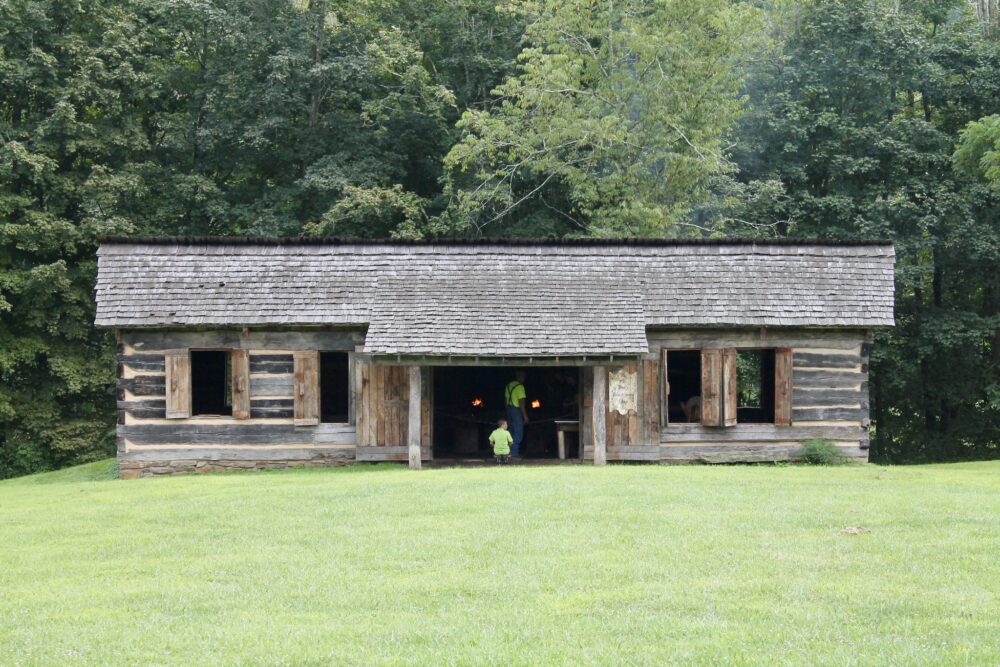 Visitors inspecting a historic wooden blacksmith house at Prickett’s Fort State Park as one of the best attractions in West Virginia, with green pasture in front and layer of trees at the back