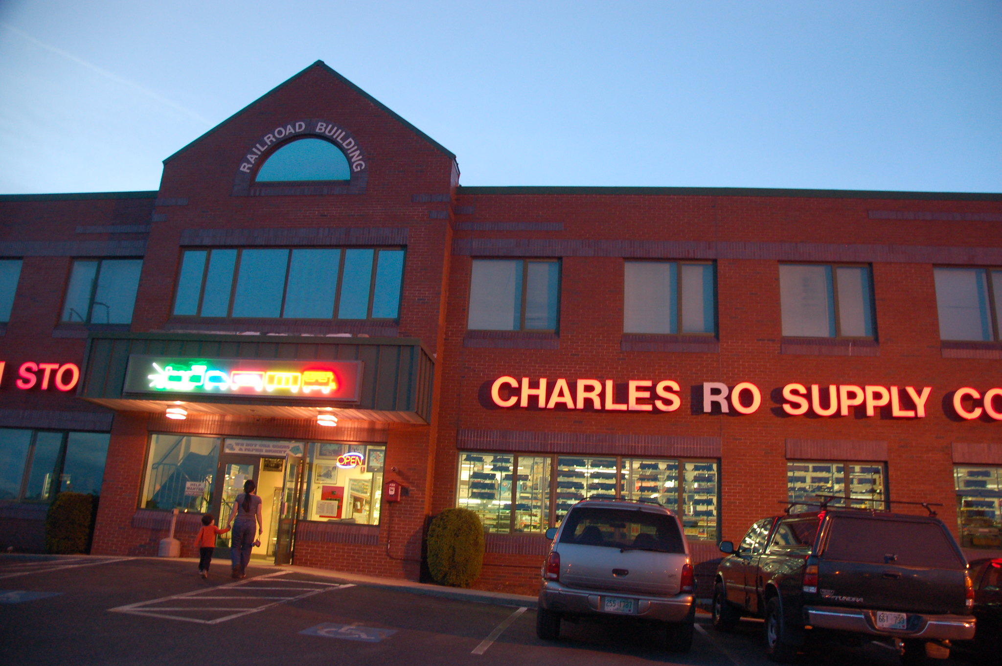 A mother with her kid heading inside one of the best toy stores in America, Charles Ro Supply Company in Malden at dusk with its signage lights turned on and cars parked outside