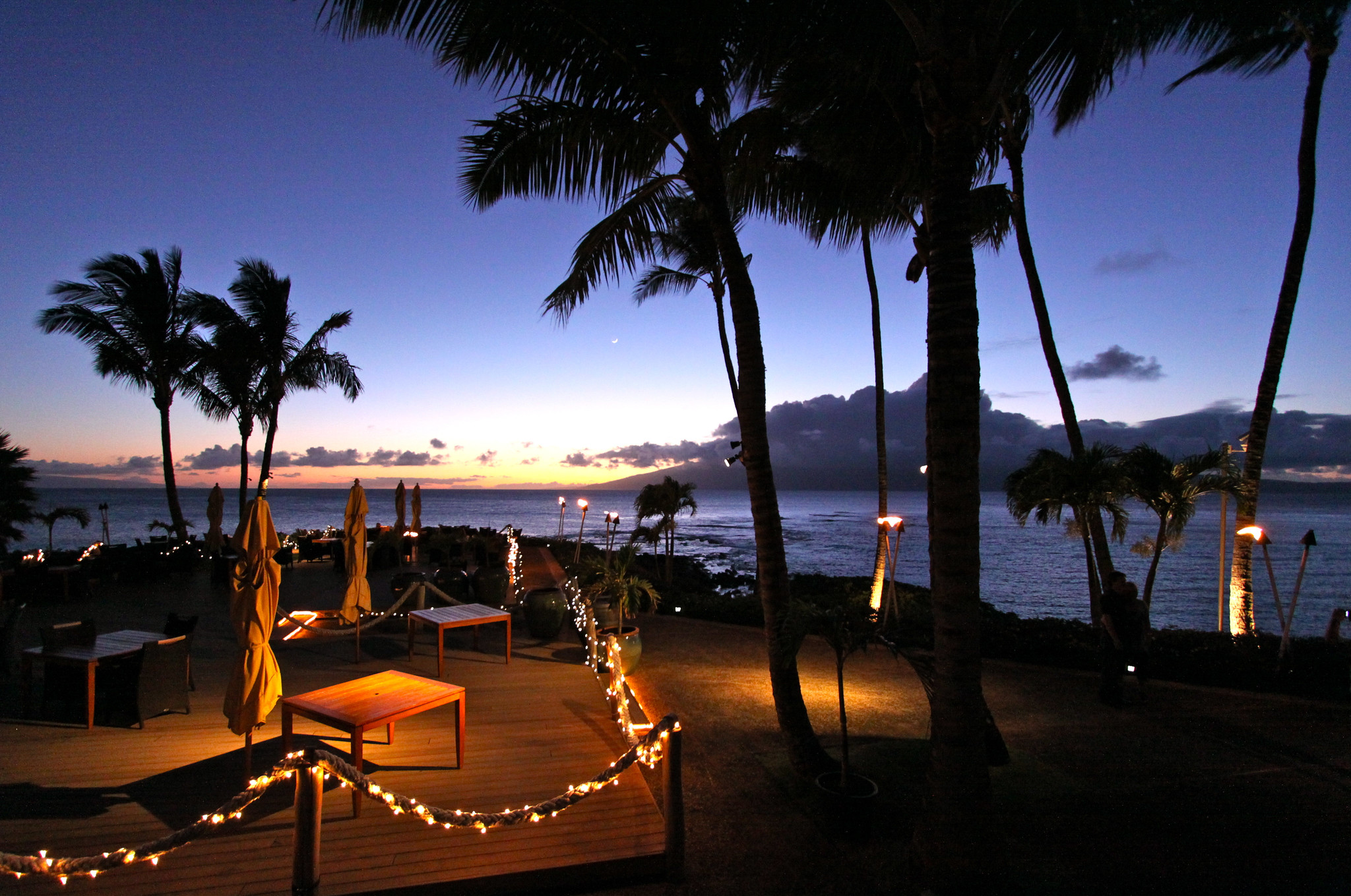 Lovely sunset view at the romantic beach-dining Merriman’s Kapalua, one of the best restaurants in Maui, Hawaii, with series lights interlaced with ropes and palm trees silhouette 