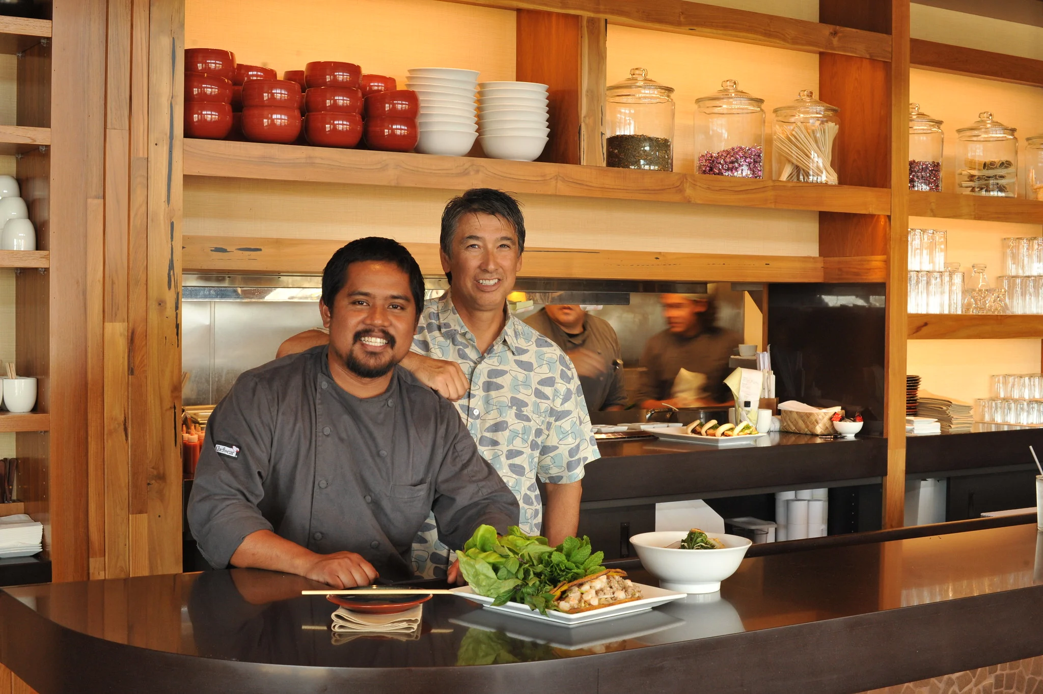 Chef Sheldon Simeon and a friend posing for a photo with a delicious food on the counter at Star Noodle noted as one of the best restaurants in Maui, Hawaii, and two chefs busy at the kitchen in background