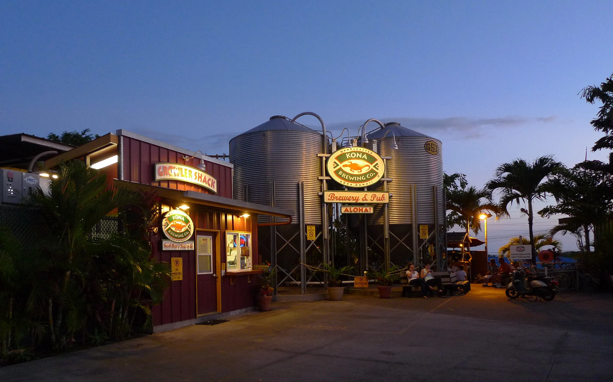 A chill dusk at Kona Brewing Company, a piece on the best restaurants in Kona, Hawaii, with lighted signs and few people on benches beside its brewing tanks
