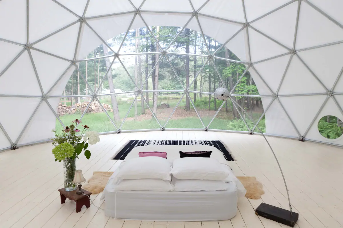 A simple interior with a single bed and a lamp at Outlier Inn Geodome in New York, with its transparent glass wall made from interconnected triangles