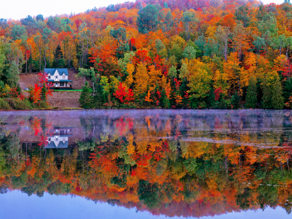 A lonely cabin with a parked car and bright colored trees on an Autumn season, reflection on the calm and clear lake at Laurentian Mountains, one of the best places to visit in Canada