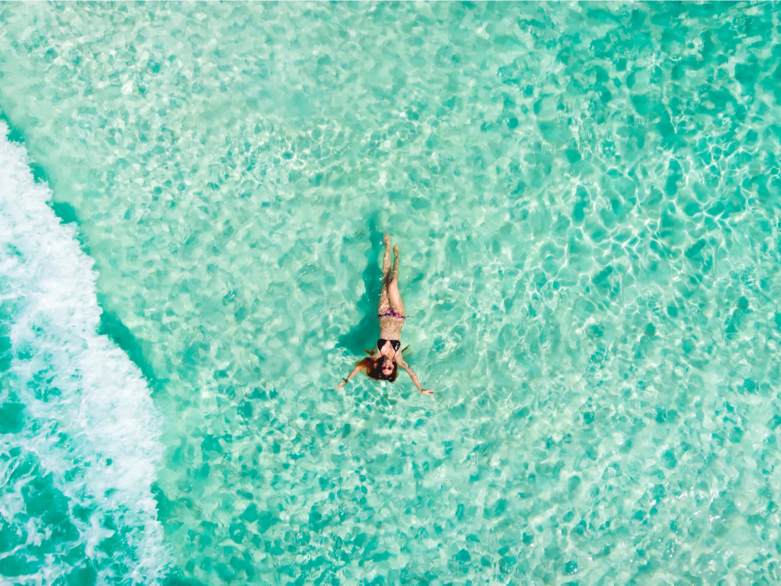 Overhead view of a young woman on her swimwear and sunglasses floating at the shallow waters in one of the best beaches in Cancun