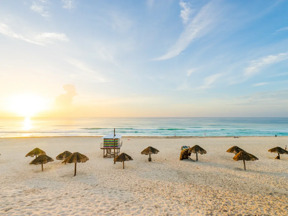 Several single pole tiki huts randomly placed on the fine shore of Playa Delfines, one of the best things to do in Cancun, on a sweet fresh sunrise with calm waves