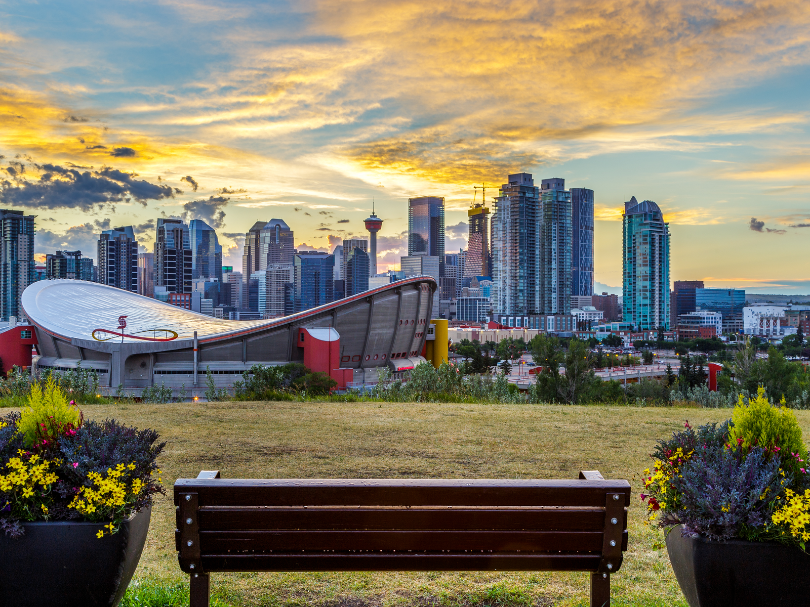 A lonely bench, sitting in between two large pots of plants, fronting Calgary City Skyline, one of the best places to visit in Canada, on a magnificent sunset