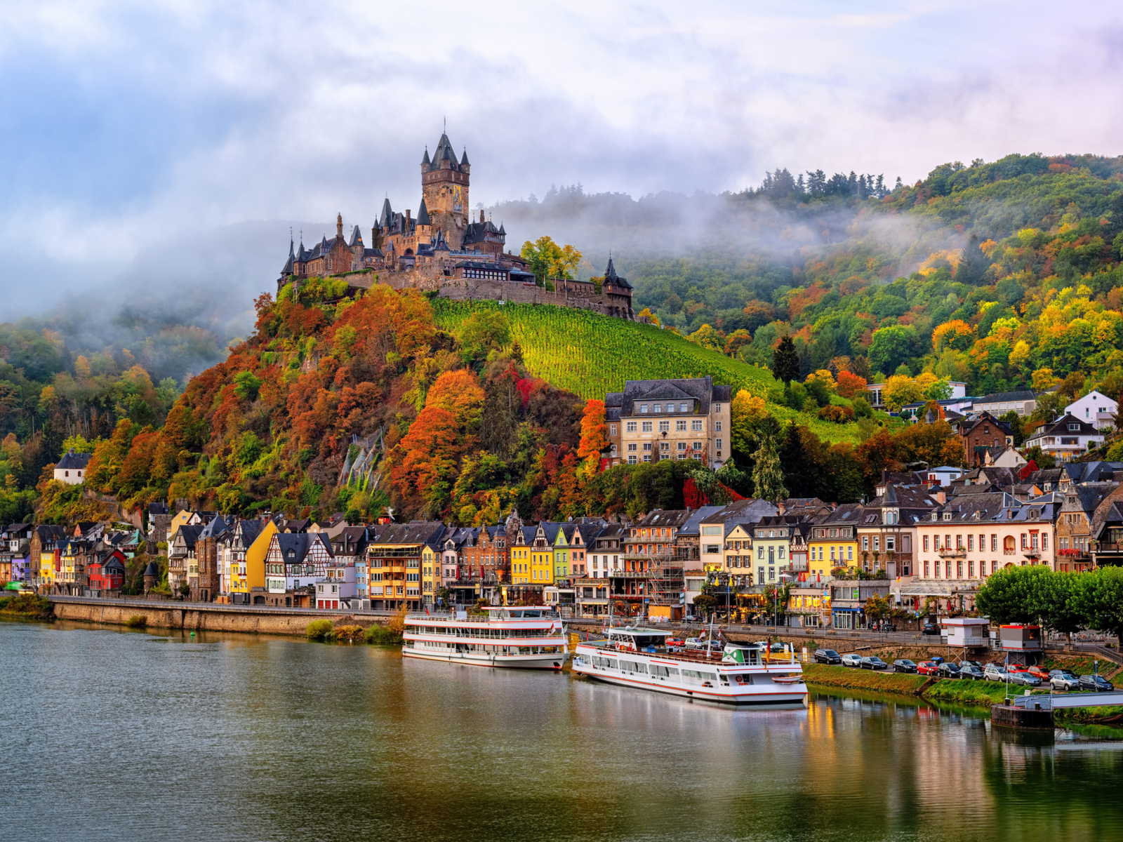 Mistry Cochem Imperial Castle also known as Reichsburg Castle is one of the best castles in Germany, erected on top of a hill at Cochem Town on Autumn with two passenger boats on the calm Moselle River