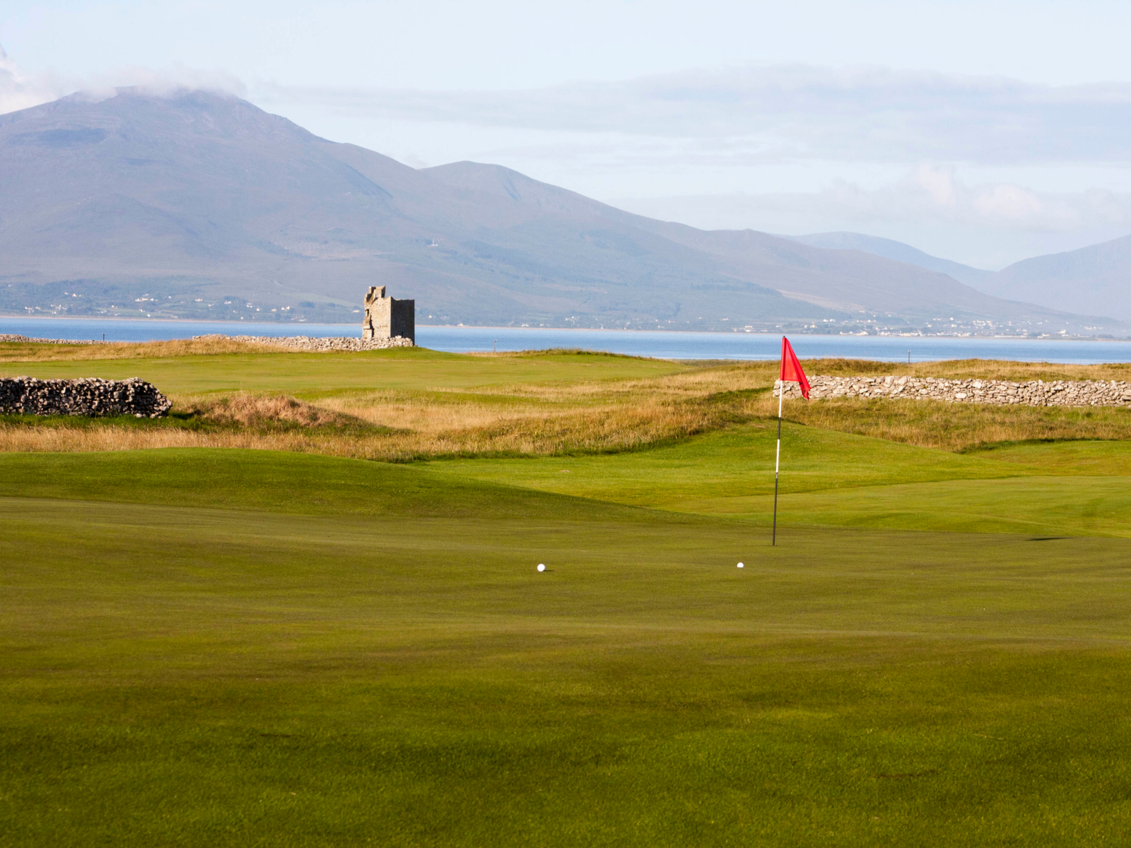 Two golf balls sitting next to a small red flag at Tralee Golf Club, a piece on the best golf courses in Ireland, with a calm sea and tall mountain in background