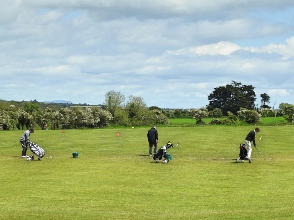 Three golfers freely striking golf balls into the vast green field at County Louth Golf Club in Baltray, one of the best golf courses in Ireland, where hundreds of white golf balls scatter at a distance