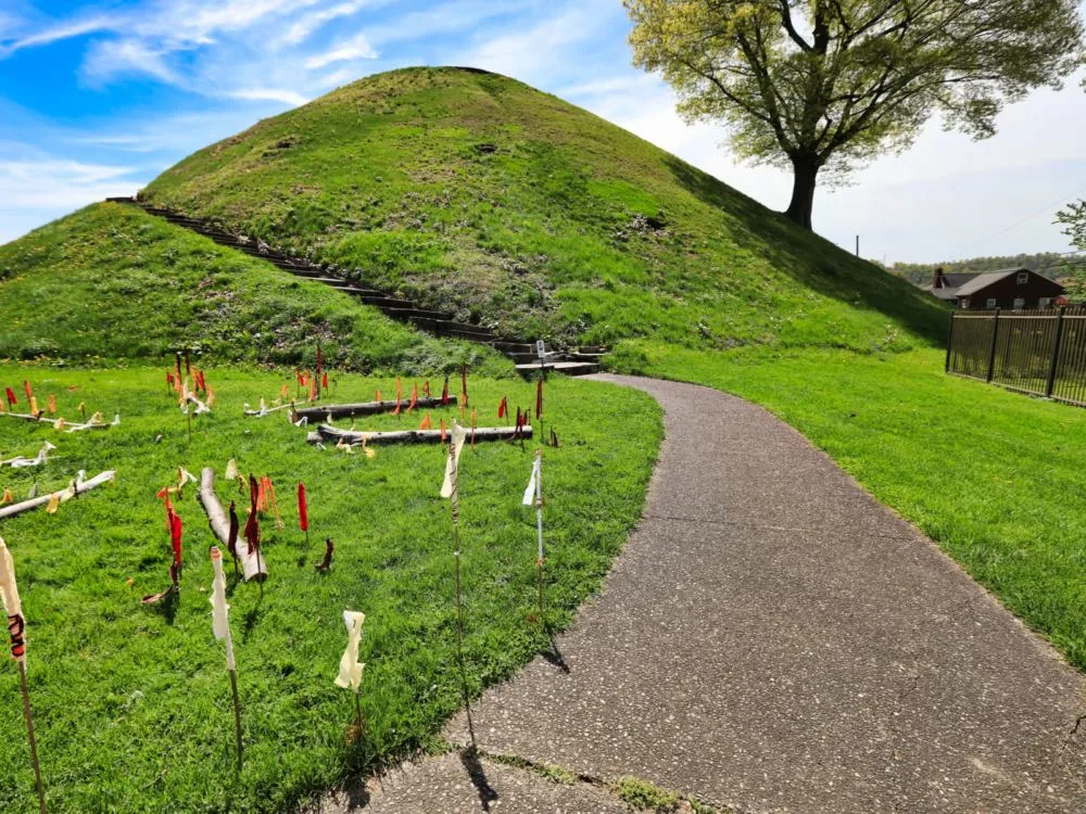 A path leading towards a mound of soil, with man-made stairs and a single tree planted on one side, is a historical burial place at Grave Creek Mound and one of the best attractions in West Virginia