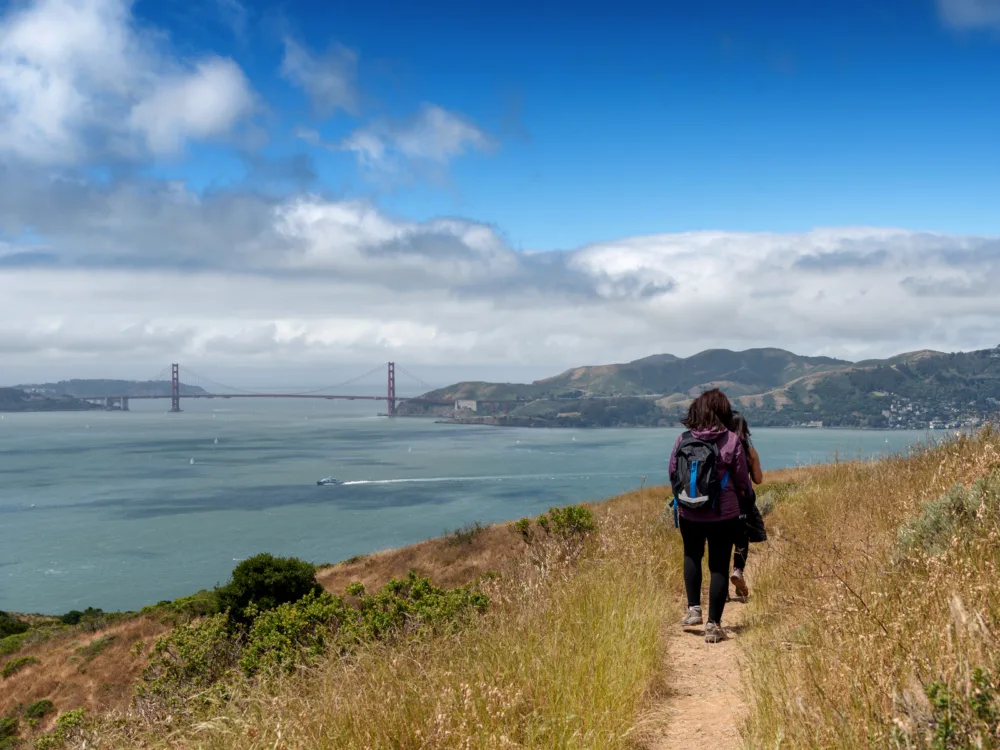 Two women trekking a a trail to Mt. Livermore on Angel Island State Park, a piece on the best places to visit in San Francisco, and a mass of cloud hovering over the Golden Gate Bridge at a distance