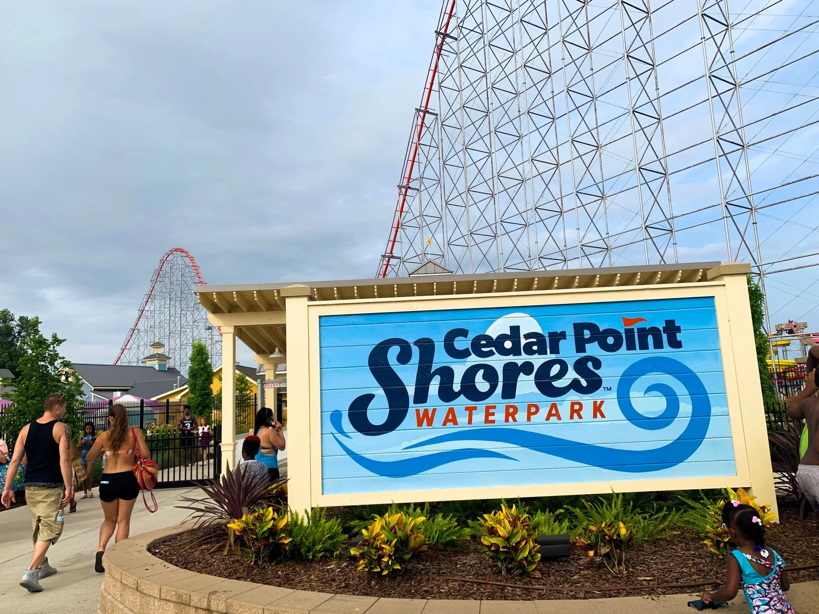 People in their summer outfit entering Cedar Point Shores Waterpark in Sandusky, OH, one of the best water parks in the USA, with a long roller coaster track in background and a child playing in front of Cedar Point Shores Waterpark signage 