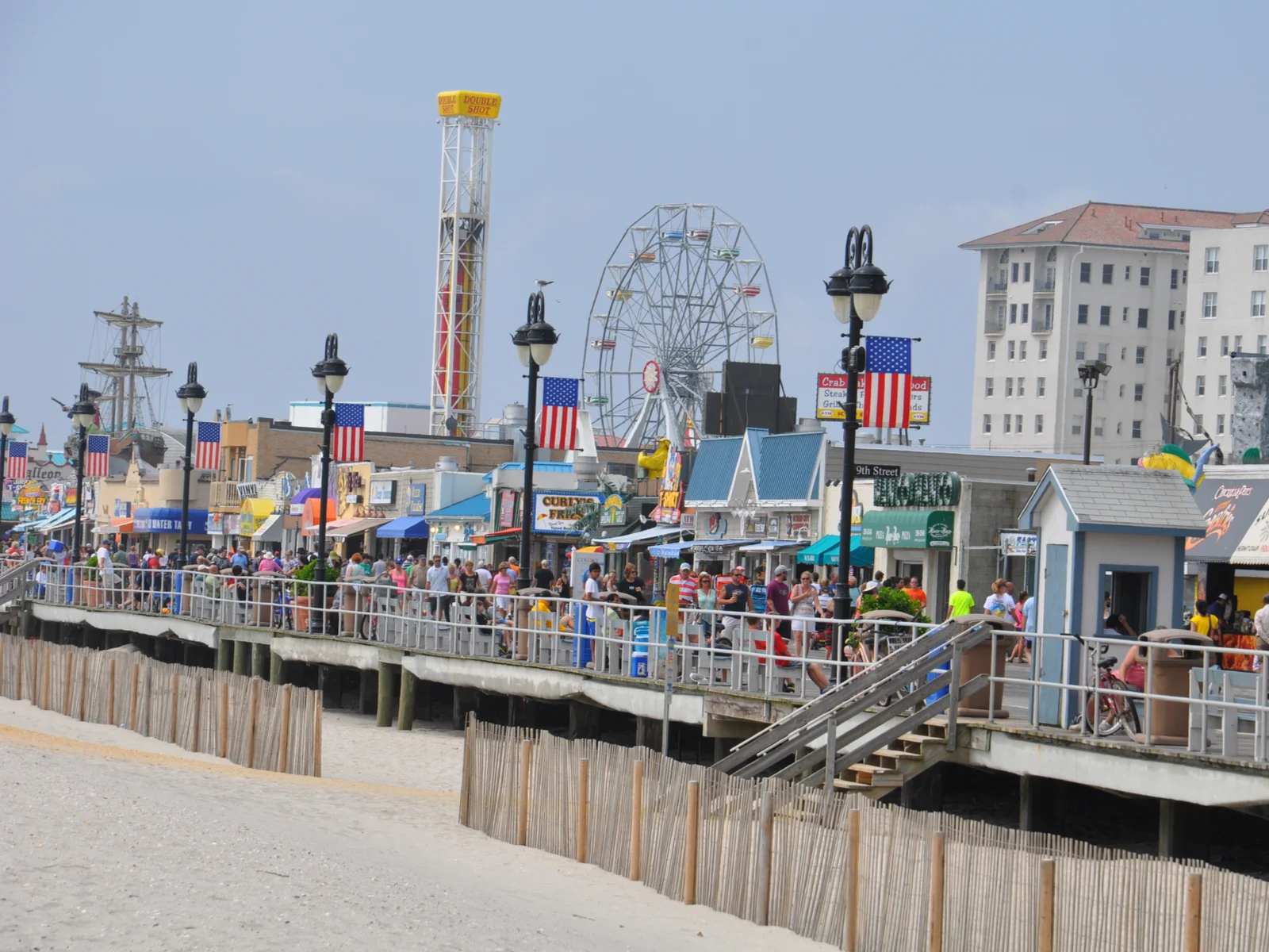 A busy day at Ocean City Boardwalk with a series of food stores, a medium-size ferris wheel and boat with sails in background, one of the best things to do in New Jersey