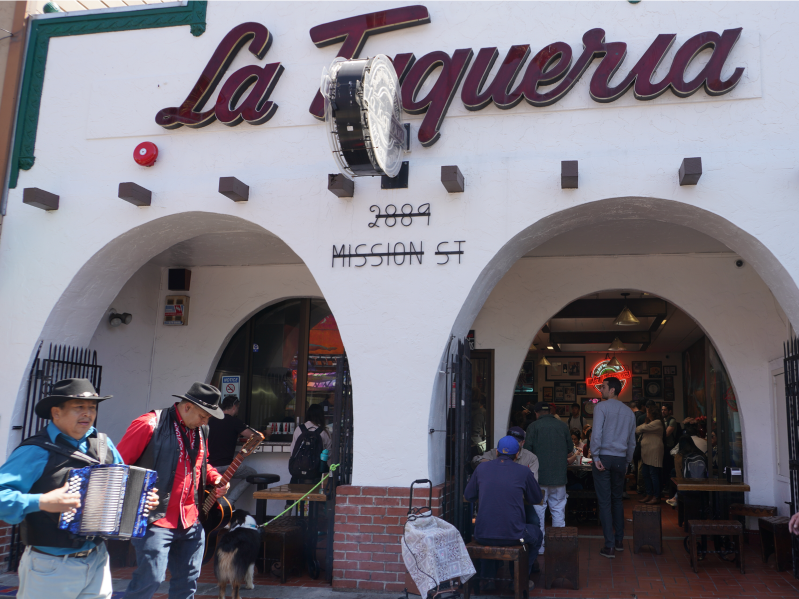Two people carrying an Acoustic Guitar and an Accordion passing by La Taqueria filled with costumers in Mission District, known to make the best Burritos and one of the best places to visit in San Francisco