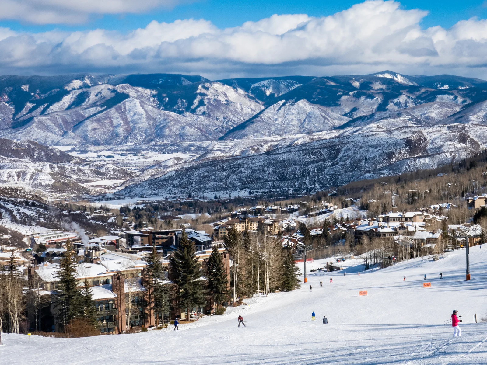 Pictured people skiing down a trail at Aspen Snowmass Ski Resort and the skyline of Rocky Mountains, a piece on one of the best ski resorts near Denver