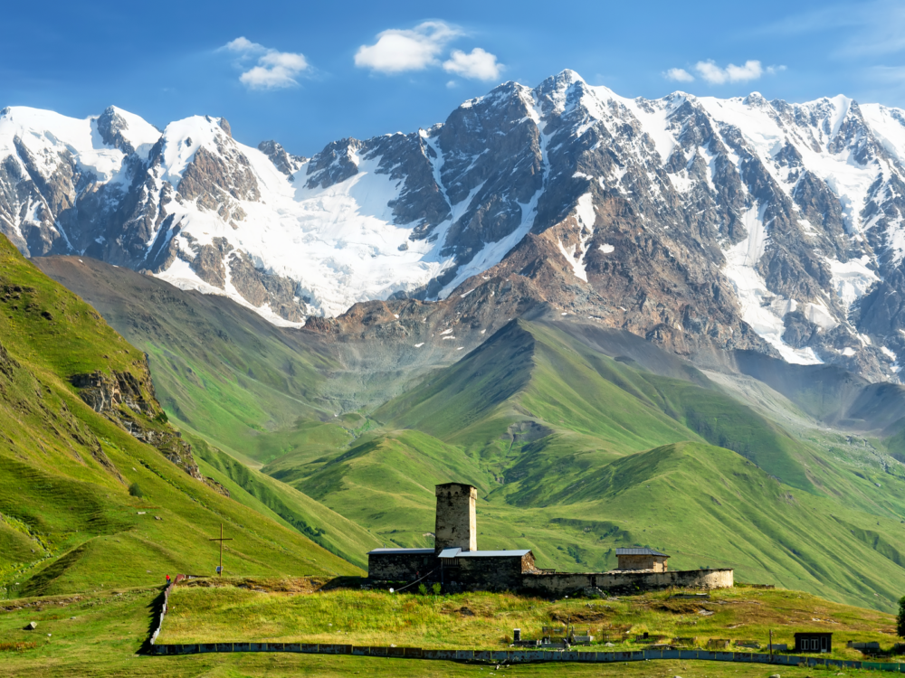 Snowy peak at Georgia's highest summit, one of the best tourist attractions in Georgia, Caucasus Mountains with green fields at its foot and Shkar and Lamaria Monastery
