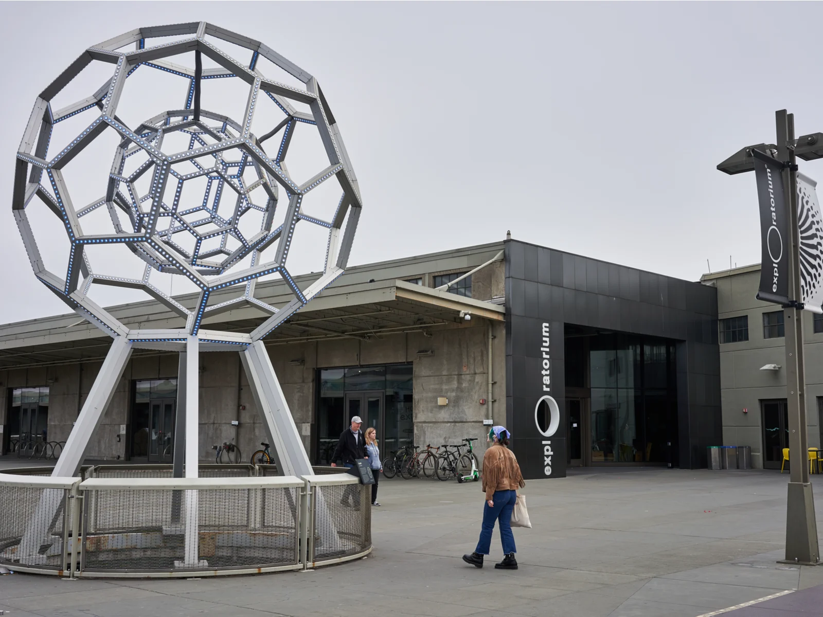 Three people walking beside a modern statue and several bicycles parked beside Exploratorium, technology and arts in San Francisco and one of the best science museums in America