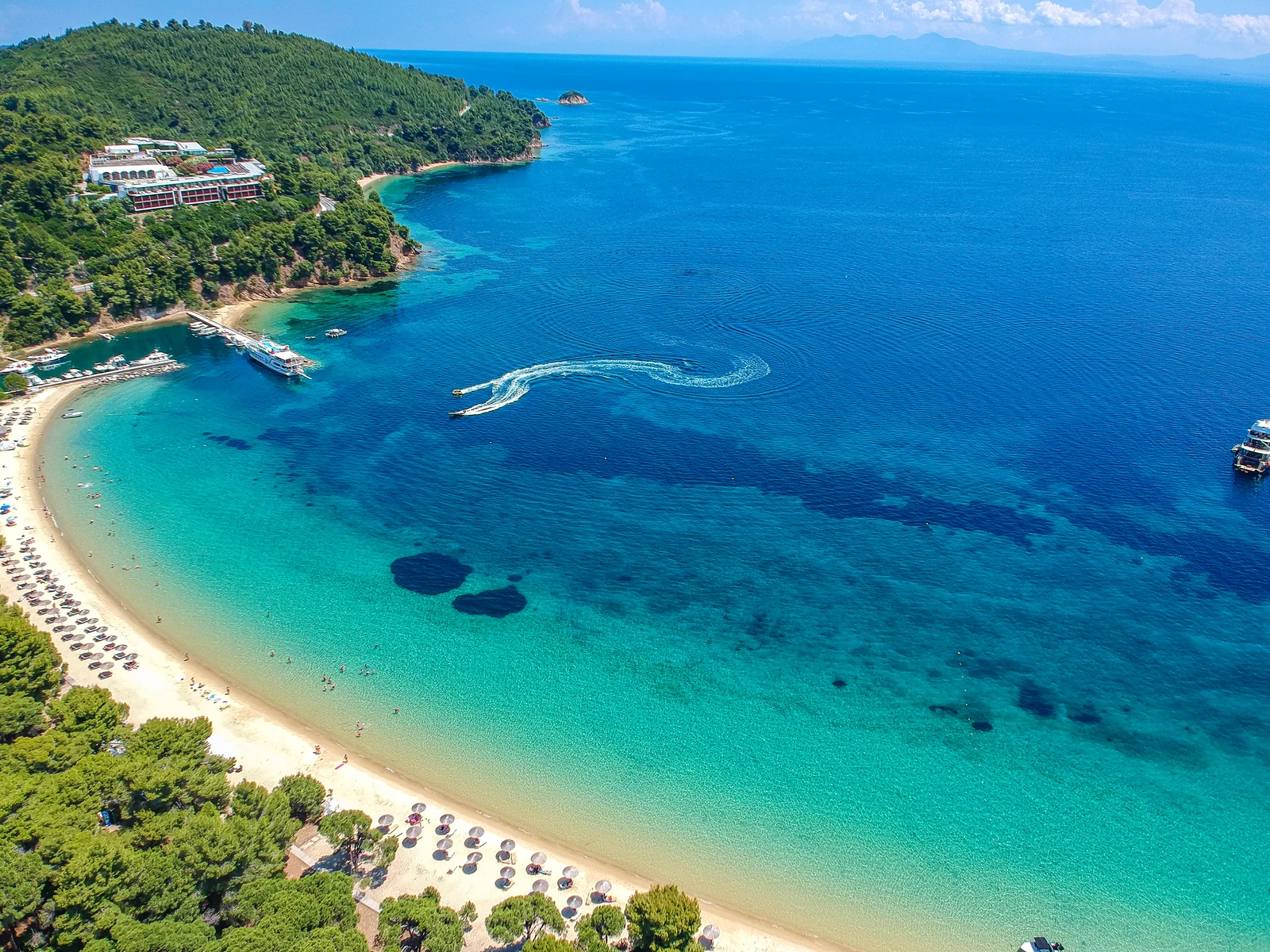 Drone view of a beautiful beach at Skiathos Island, one of the best islands in Greece to visit, where sunloungers are perfectly lined on shore, few docked and sailing ships, and tourists enjoying a banana boat ride on crystal clear water