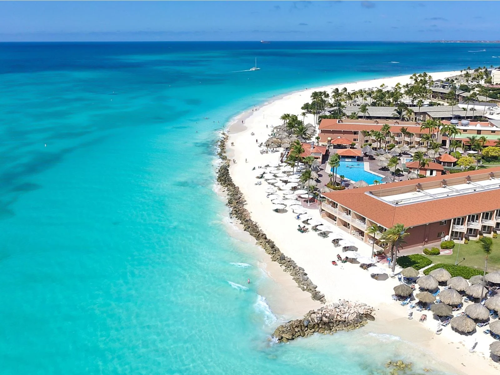 Large hotels, several native huts, tall palm trees, tourists on sunloungers with white umbrellas and others enjoying the beach and shore, and boats sailing at the distant deep waters of Manchebo Beach, seen on an aerial view at one of the best beaches in Aruba