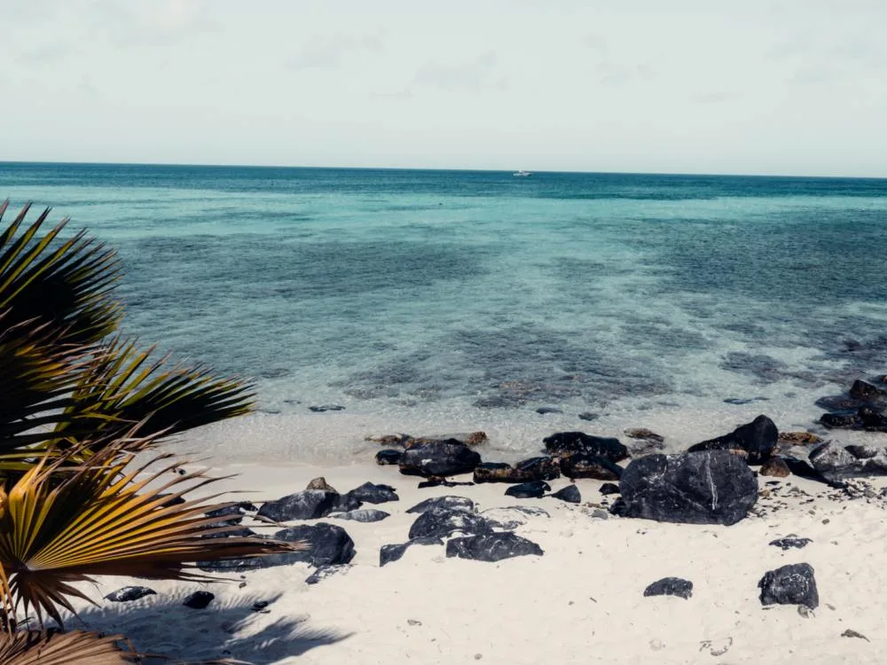 Large rocks, fine sand, clear waters, and a ship at a distant on Malmok Beach, photographed as a piece on the best beaches in Aruba