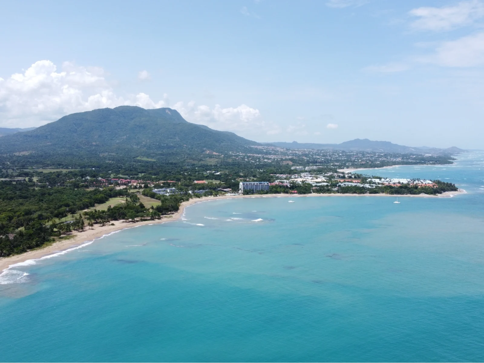 Aerial view of the winding shoreline and calm clear water of Playa Dorada in Puerto Plata, one of the best beaches in the Dominican Republic with a massive mountain and a populated region 