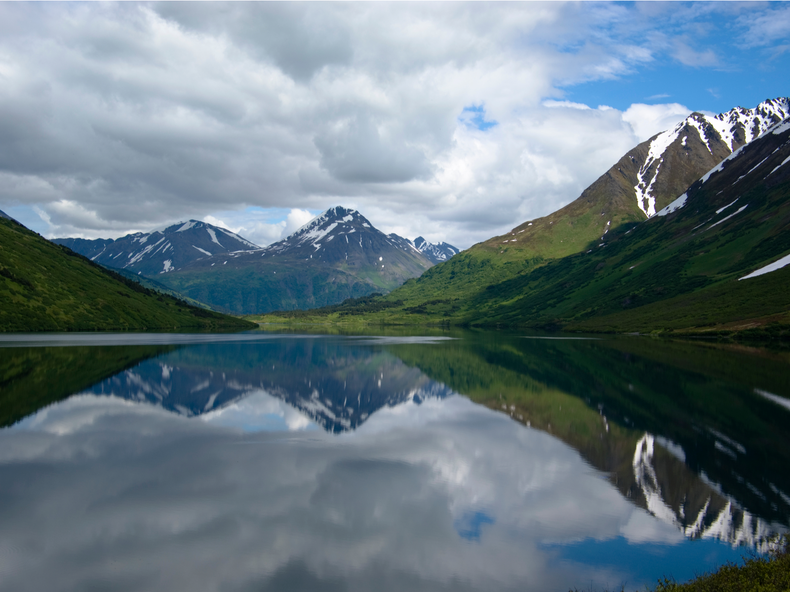 Thick white clouds and green snowy peak mountains reflected on the still water of Crescent Lake in Alaska, one of the best lakes in the U.S.