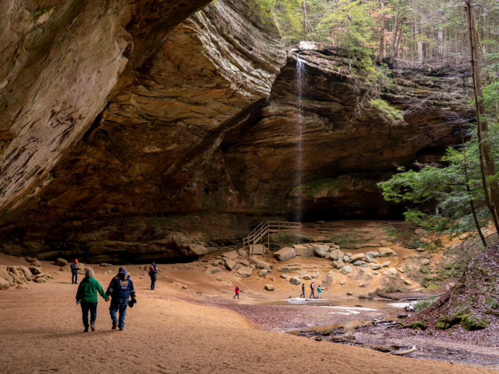 A couple tourist, holding each other's hands as a piece on the best things to do in Ohio, and company walking underneath Ash Cave in Hocking Hills State Park