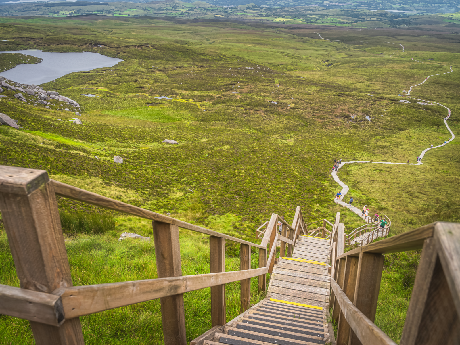 People climbing down the long winding boardwalk trail circled about by green pasture at Cuilcagh Legnabrocky Trail, one of the best hikes in Ireland