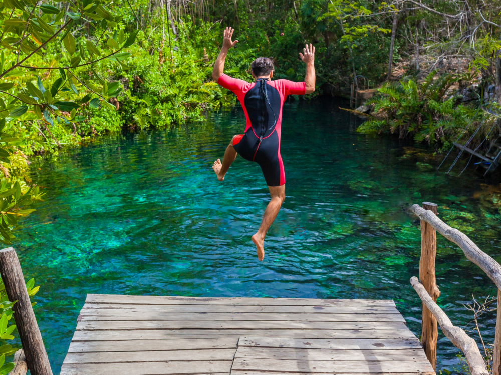 A man on his rash guard animatedly jumping off a boardwalk to the crystal clear water of Riviera Maya, one of the best things to do in Cancun, at Tulum