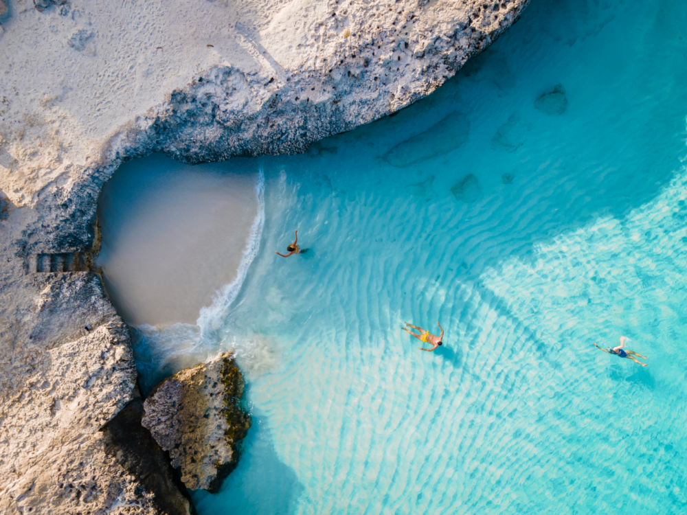 Overhead view of three people swimming on the crystal clear calm waters of a rocky coast in Triple Steps Beach, one of the best beaches in Aruba, during a bright day