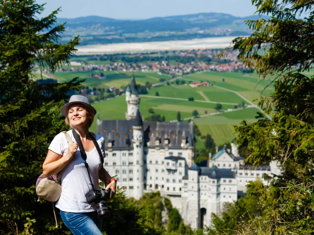 A smiling woman holding her camera with the famous Neuschwanstein Castle blurred in background, one of the best castles in Germany
