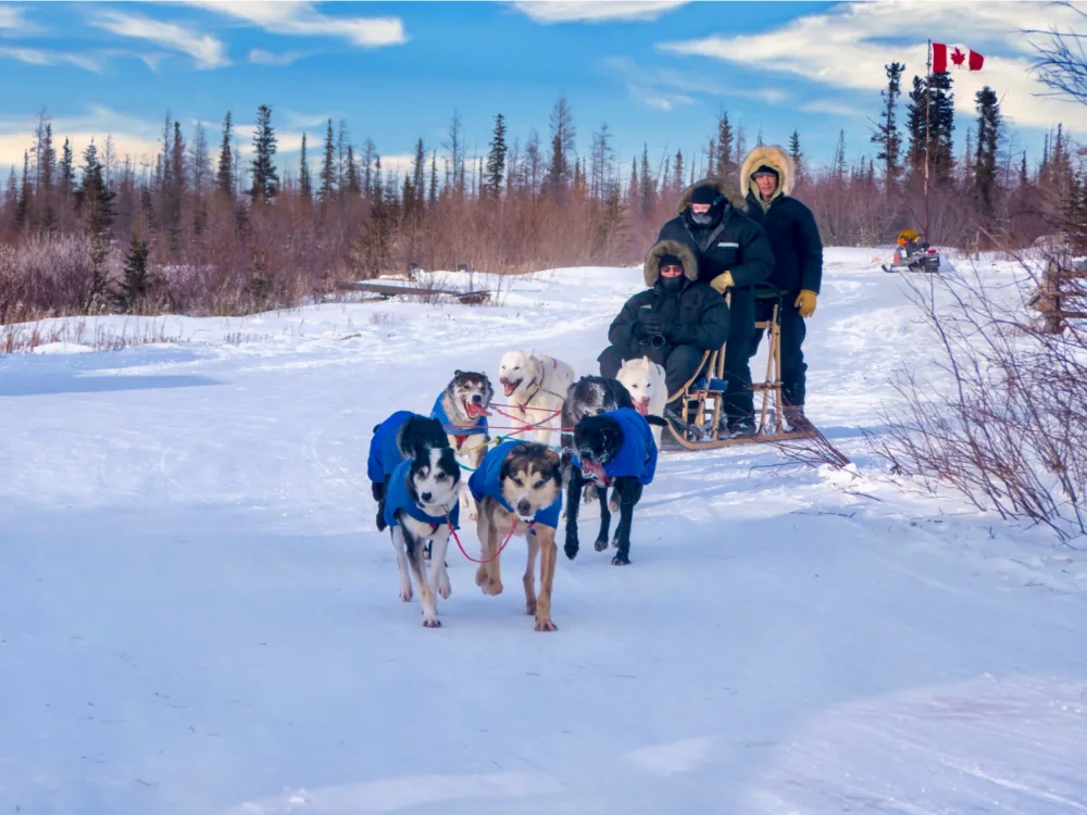 Three people in their winter gears riding a traditional dog sled being pulled by a team of six huskies in Churchill, Manitoba, one of the best places to visit in Canada, on a freezing winter