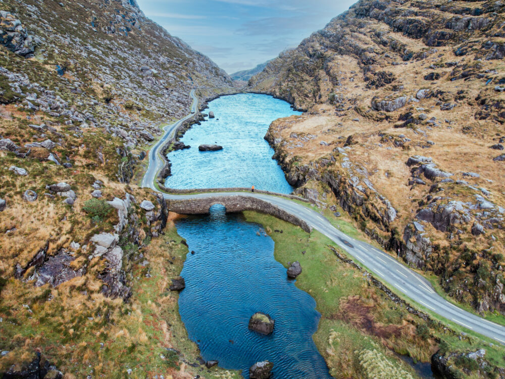 Aerial view on stone Wishing Bridge crossing a tranquil stream, and winding road on the side of rocky valley at Gap of Dunloe, one of the best hikes in Ireland