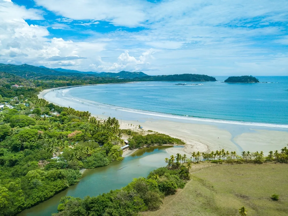 Wide view of Playa Samara and its river estuary emptying into the ocean on a nice day with cloud cover for a guide showing the top beaches in Costa Rica