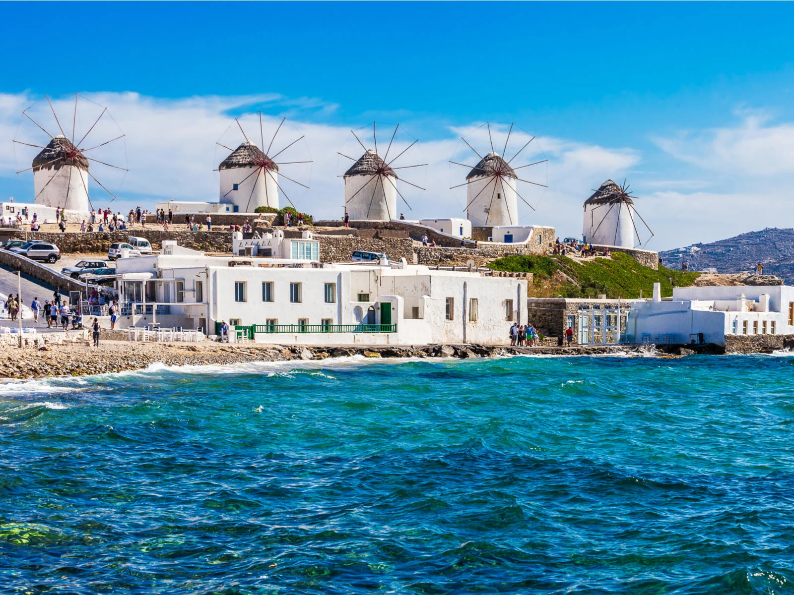 Tourists gathered around the five iconic white windmills at Mykonos, photographed from the the wavy blue see as a piece on the best islands in Greece to visit