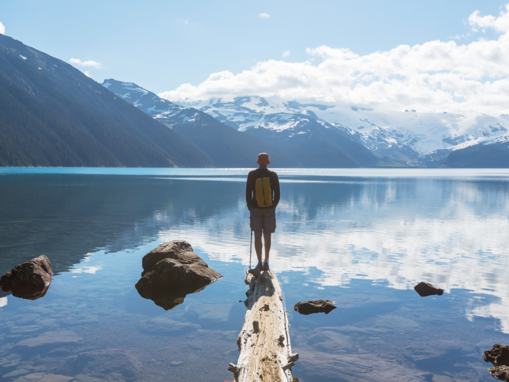 A man with a walking stick standing on a drift wood facing the crystal clear Garibaldi Lake, one of the best places to visit in Canada, near Whistler, British Columbia 