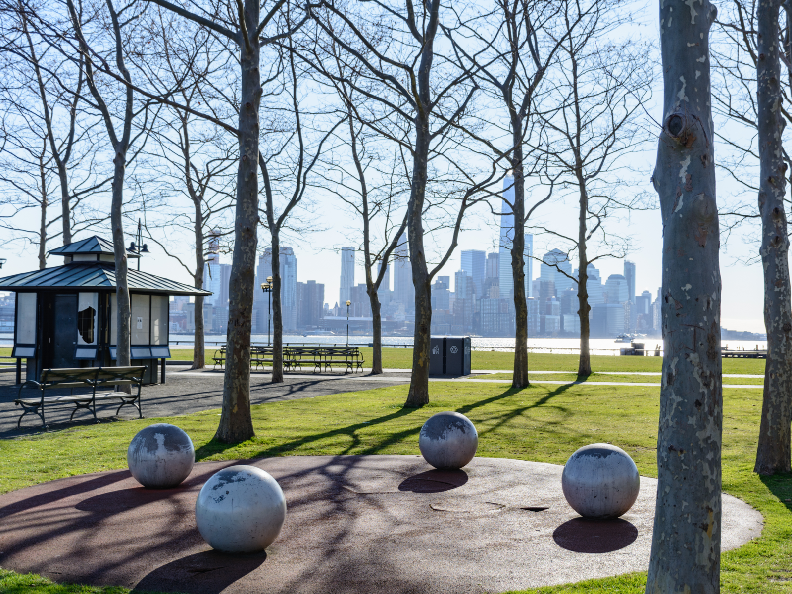 Four spherical objects, bare trees, empty benches, a small store stall, and a view of New York City at Liberty State Park during a fall season, having a picnic here is one of the best things to do in New Jersey