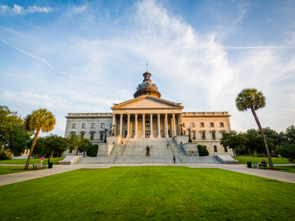 A milky sky over a wide angle view on South Carolina State House, considered as one of the best South Carolina attractions, fronting a wide green lawn where people seen strolling