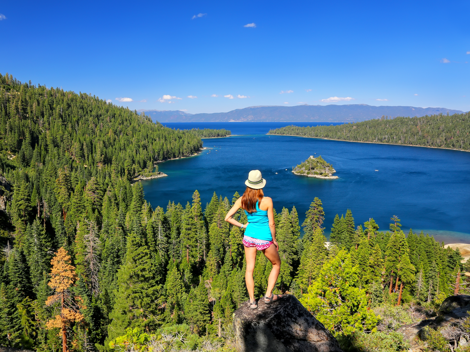 A young woman on her sun hat standing on rock overlooking the lush pine tree forest and peaceful Emerald Bay at Lake Tahoe in California and Nevada, one of the best lakes in the U.S.