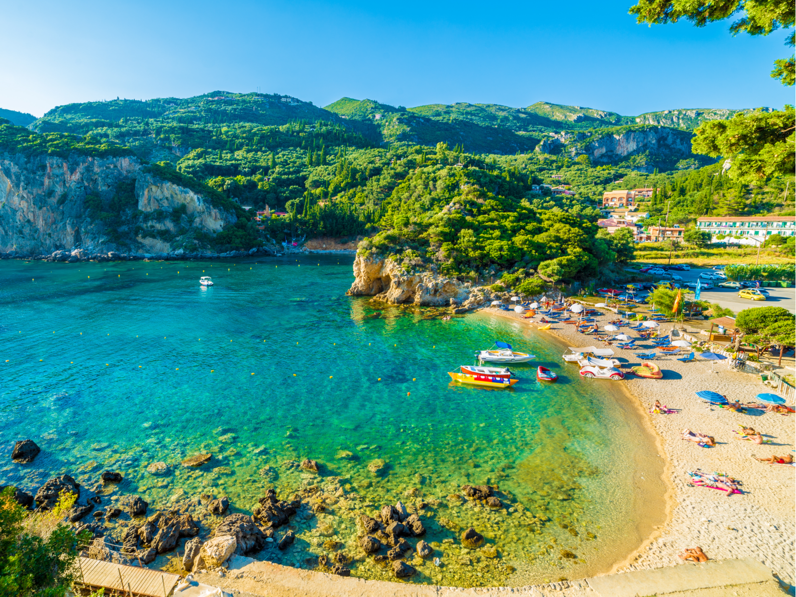 Tourists sunbathing on a beautiful beach during a summer at Corfu Island, a piece on the best places to visit in Greece, and small boats idling on the calm emerald waters